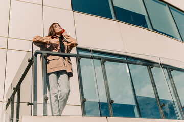 Caucasian young pretty stylish woman standing near business office center glass balcony railing dressed brown trench coat smiling happy outside in city park, spring autumn season. Brunette hair lady