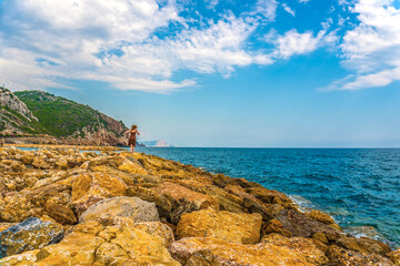 Fototapeta na wymiar Young woman or girl on a rocky seashore wearing dress and straw hat in Alanya resort, Turkey