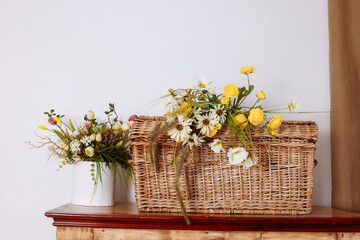 A wicker basket filled with wild flowers stands on a wooden table against a white wall.