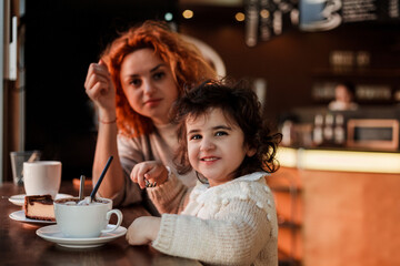 Beautiful young red-haired mother with cute curly-haired daughter are sitting in cozy cafe and drinking hot school. Mothers Day. Warm.