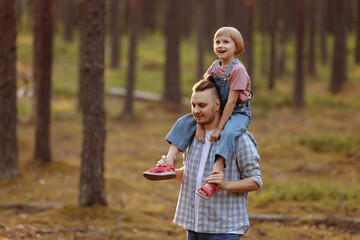 Dad holds a little daughter in his arms, they laugh merrily walking along a forest road among tall pines. Family walk in the forest at sunset, man and little girl. Baby sits on dad's shoulders.