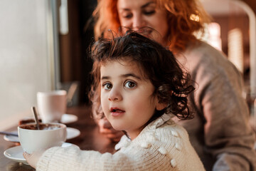Beautiful young red-haired mother with cute curly-haired daughter are sitting in cozy cafe and...