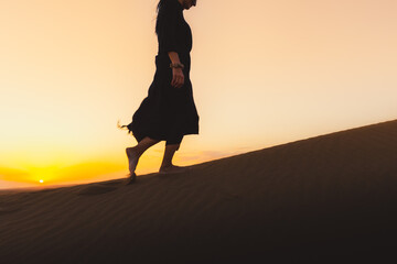Woman black dress walking in the Dubai desert sand dunes with footsteps in the sand during sunset,...