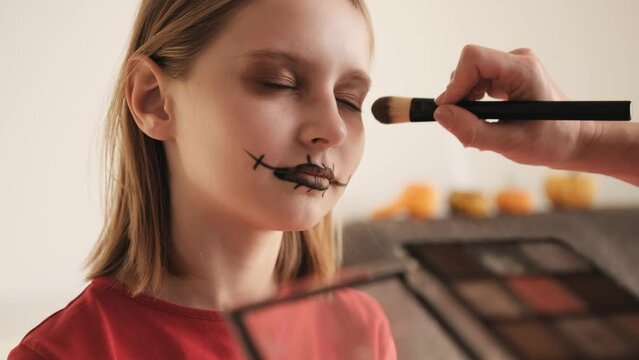 Mother making Halloween makeup to preteen girl daughter and painting spooky black lines on face. Pretty child with creepy cosmetics preparing for October holiday party