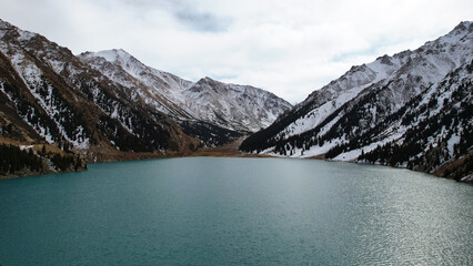 Obraz premium A lake in the mountains with turquoise blue water. Drone view of clear water, coniferous trees and snowy mountains. People walk along the shore, low bushes grow. Big Almaty lake. Kazakhstan