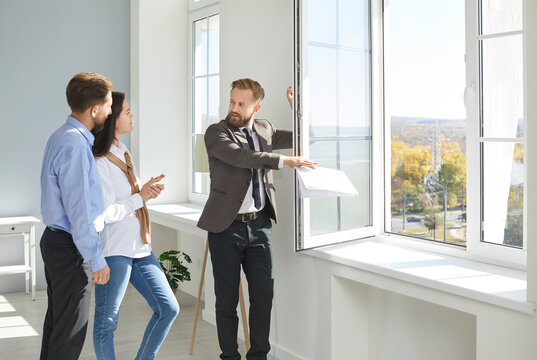 Real estate agent shows new home. Male realtor shows apartment for sale and its view from window to young married couple. Family looking for house to buy listens to man in suit. Real estate concept.