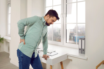 Handsome oung man suffering from backache in ofiice. Office worker standing leaning table touching...