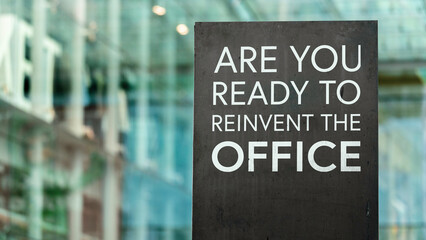 Are you ready to reinvent the office on a black city-center sign in front of a modern office building	
