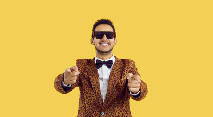 Happy cheerful ethnic black man in stylish glasses, extravagant leopard jacket and bowtie isolated on yellow background pointing both index fingers at camera, smiling and inviting you to fun party