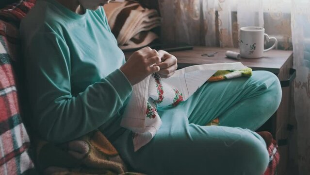 Young woman sews manually with thread and needle in slow motion. Woman in casual home clothes embroidering while sitting on a cozy sofa at home. Warm atmosphere, natural daylight, lifestyle, handmade