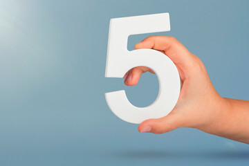 Number five in hand. Hand holding white number 5 on blue background with copy space. Concept with number five. Birthday 5 years, fifth grade, five day work week - obrazy, fototapety, plakaty