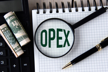 Letters OPEX under a magnifying glass on a white notebook against the background of a fragment of a calculator and banknotes