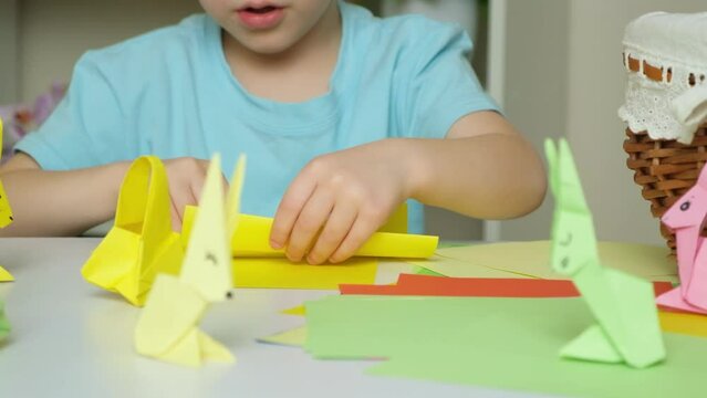 Preschool boy with rabbit ears nag head makes Easter crafts, origami figurines of rabbits and baskets of paper