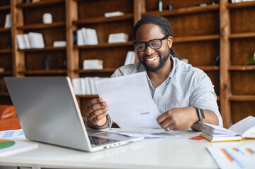 Smiling positive african-american male male employee, entrepreneur or businessman holding document, bill or contract, man reading great news in letter received sitting at the office desk