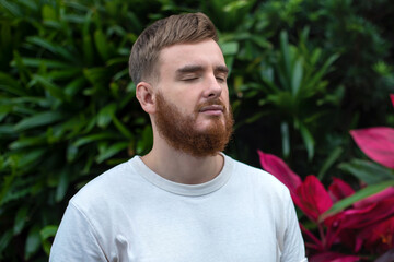 Portrait of happy young bearded man with beard breathing deep, inhale fresh air on natural background, green wall with leaves, flower in tropical country in garden. Eco, ecological concept. 