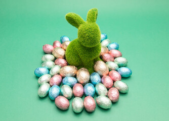 Green bunny, rabbit surrounded by small Easter chocolate eggs, sweets wrapped in colorful bright...