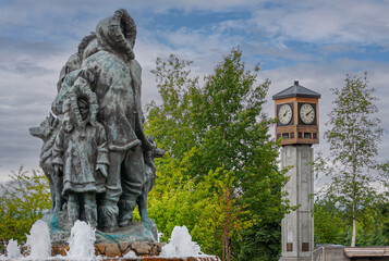 Fairbanks, Alaska, USA - July 27, 2011: Unknown First Family statue and fountain on Golden Heart Plaza. Child and father side, with Rotary Clock Tower and green foliage under blue cloudscape - obrazy, fototapety, plakaty