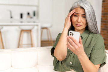 Frustrated unhappy gray-haired mature woman sitting on sofa, holding smartphone, senior female has not enough money for online transaction, financial problem