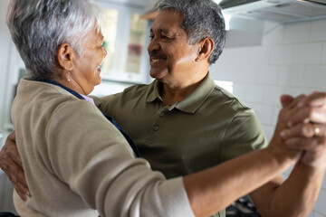 Cheerful biracial senior couple holding hands and looking at each other while dancing in kitchen