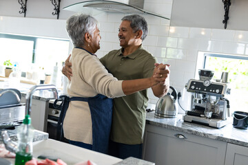Happy biracial senior couple holding hands and looking at each other while dancing in kitchen