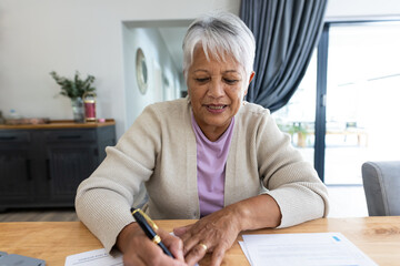Fototapeta Portrait of biracial senior woman analyzing bills on wooden table while sitting at home obraz