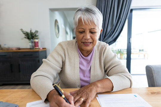 Portrait of biracial senior woman analyzing bills on wooden table while sitting at home