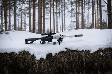 Side view of a sniper rifle with a silencer and winter camouflage in a snowy forest.