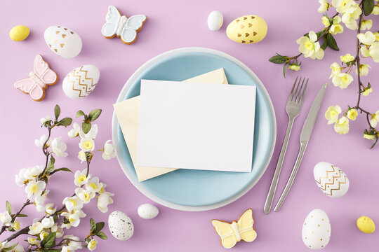 Easter celebration concept. Flat lay photo of envelope with white card on blue plate colorful eggs fork knife butterfly cookies cherry blossom branch on pastel lilac background