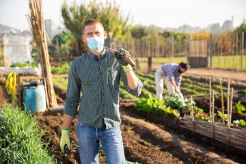Portrait of confident male gardener in protective face mask standing with shovel during spring...