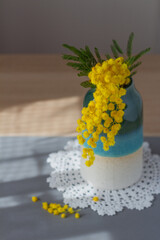Yellow spring flowers in a vase on a white napkin and wooden table, a sprig of mimosa, light and shadow, close-up.