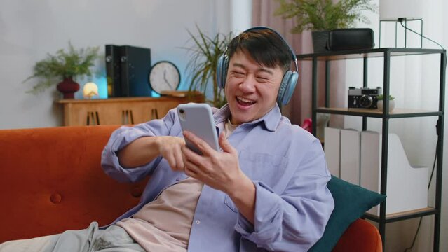 Happy overjoyed asian man in wireless headphones relaxing sitting on couch at home apartment choosing listening favorite energetic disco dancing music. People weekend daytime leisure activities