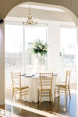 A beautiful flower bouquet centerpiece with white flowers and greenery sits in the middle of a wedding reception table, surrounded by gold chairs.