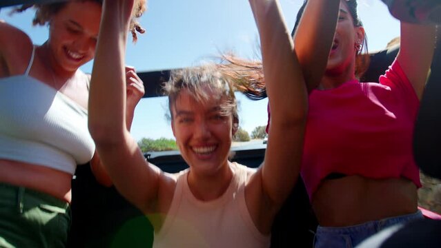 Portrait of female friends standing up through sunroof of car laughing on road trip through countryside with friends - shot in slow motion