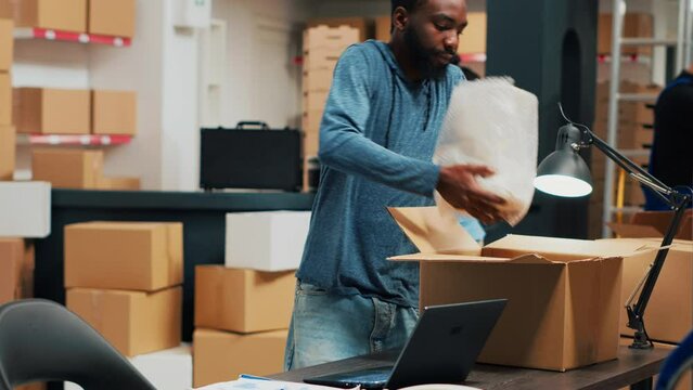 Male entrepreneur analyzing boxes with products to ship order to customers, working on stock distribution at desk. Man planning quality control and taking notes on laptop, supply chain. Tripod shot.