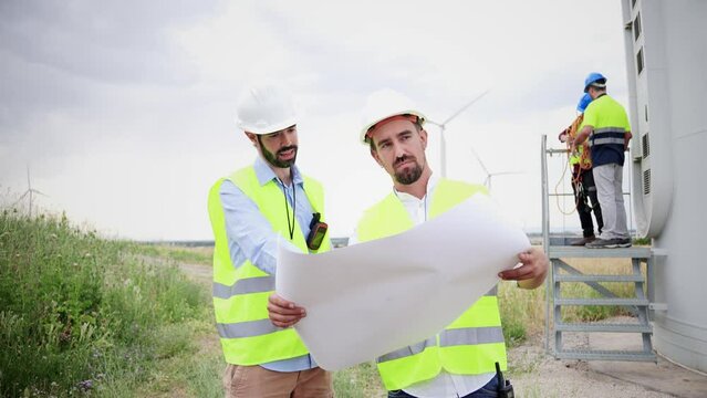 Two windmill engineers looking a blueprint with the plan of the construction of the electricy turbine. At background workers or contractors doing their job . Renewable energy concept. Slow motion