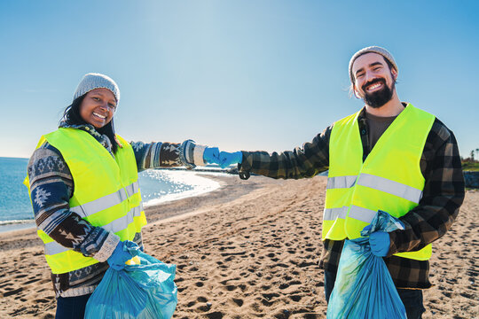 Two multiracial volunteer bumping fists after picking up garbage and plastic dirt from the sand on the beach. A couple of environmental activists smiling standing looking at camera. Ecology concept - Powered by Adobe