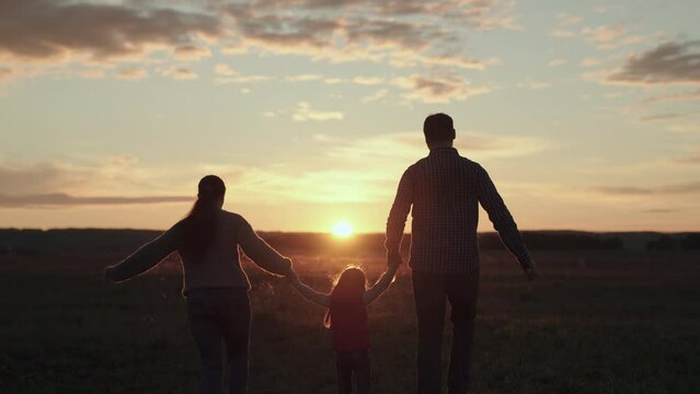 Mom, dad and daughter are playing on field, child is jumping on green grass and holding hands of parents. Teamwork. Happy family runs in park holding hands in summer at sunset. Happy healthy childhood