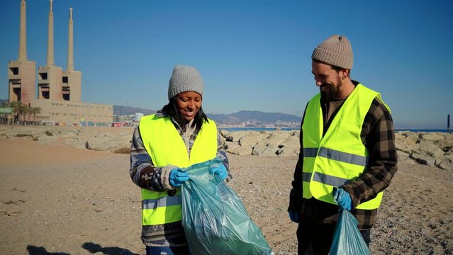 Two multiracial volunteer bumping fists after picking up garbage and plastic dirt from the sand on the beach. A couple of environmental activists smiling standing looking at camera. Ecology concept - Powered by Adobe