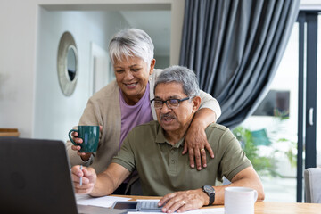 Biracial senior woman with coffee mug standing by husband and analyzing bills on table at home