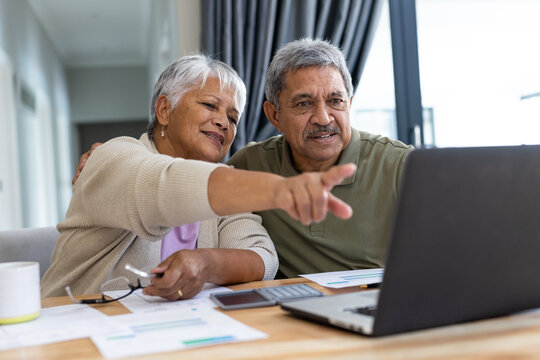 Biracial senior woman pointing at laptop and analyzing bills with husband on table at home