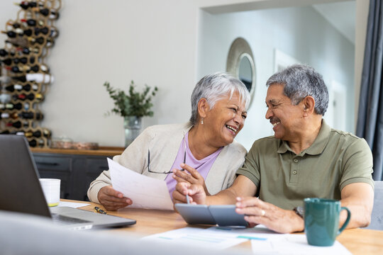 Cheerful biracial senior couple looking at each other while analyzing bills on table at home