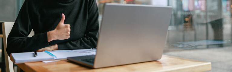 Smiling female student studying on laptop while sitting in cafe and showing thumb up