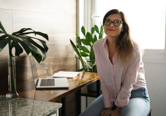 Pretty middle-aged woman wearing eyeglasses with long straight hair working at home office smiling...