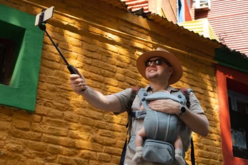 Poster Caucasian happy man, father carrying little newborn baby in carrier,family traveling, going sightseeing in center of La Boca quarter, Buenos Aires, Argentine, taking selfie photo with stick smiling. © Ольга Смолина