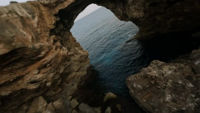 FPV drone shot of steep cliffs and natural stone bridge along the seaside in Cala Varques, Mallorca, Spain during evening time.