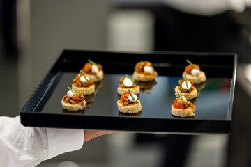 The waiter holds a Set of festive appetizer canapes on bread on a black tray as an event dish....