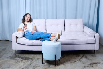 a girl in blue jeans and a white t-shirt looks at the phone while sitting on a light-colored sofa