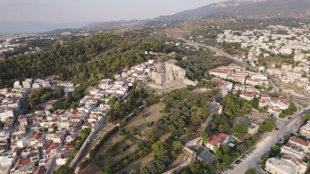 Wide circling shot of Patras Castle on a forested hilltop, Cityscape of Patras sprawling in the distance