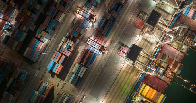 Aerial drone time-lapse top view of cargo vessel ship loading and unloading in deep sea port at night. Industrial shipyard with shipping containers being loaded onto a freight for import export.