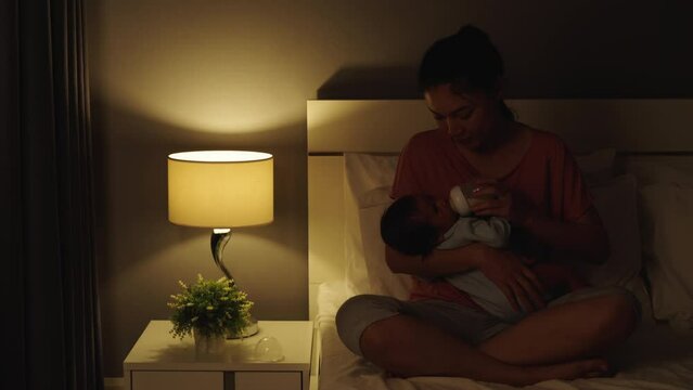 stressed mother trying to feeding milk bottle to her crying newborn baby on a bed at night